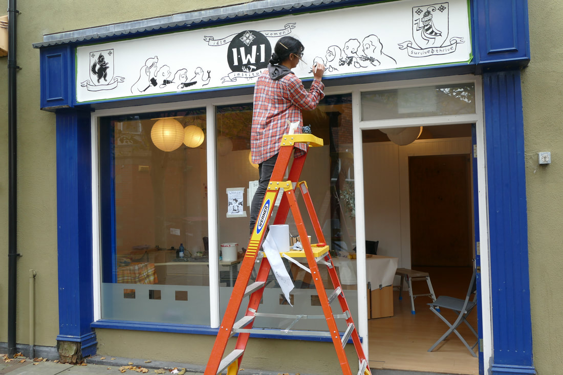 a woman at the top of a ladder painting an exterior shop sign that says IWI