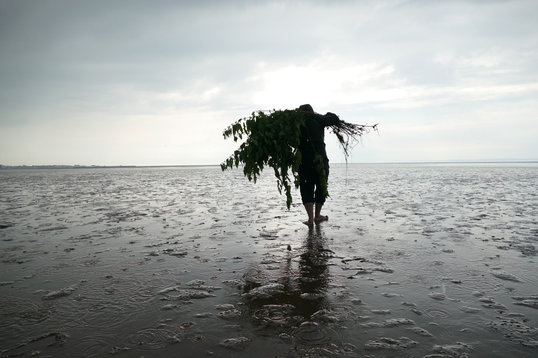 a figure balancing a large bundle of stinging nettles stands in the rain on a vast grey beach